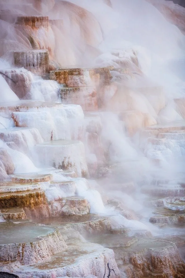Terraced hot springs cascading down a hillside, with mineral deposits creating a white and rusty rim around each pool, and steam rising from the water.