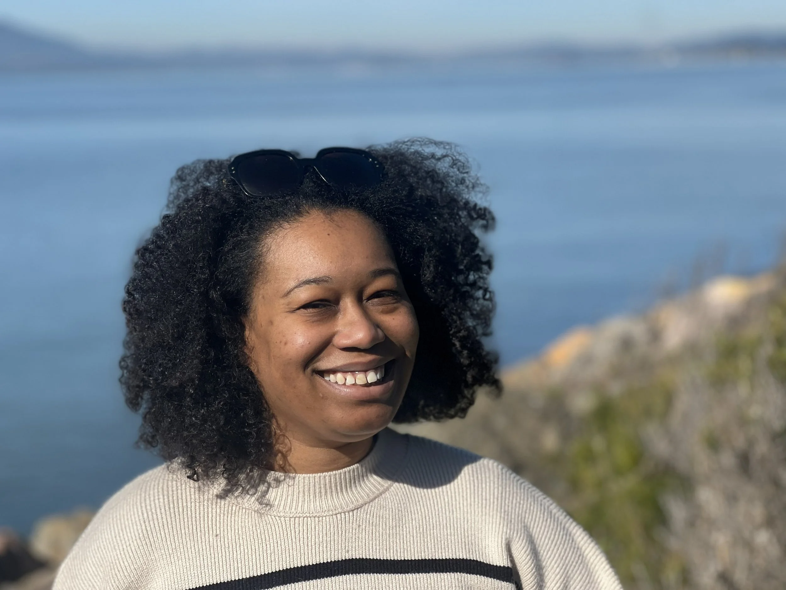 Smiling woman wearing a white and black top, in the sun, in front of rocks and water. 