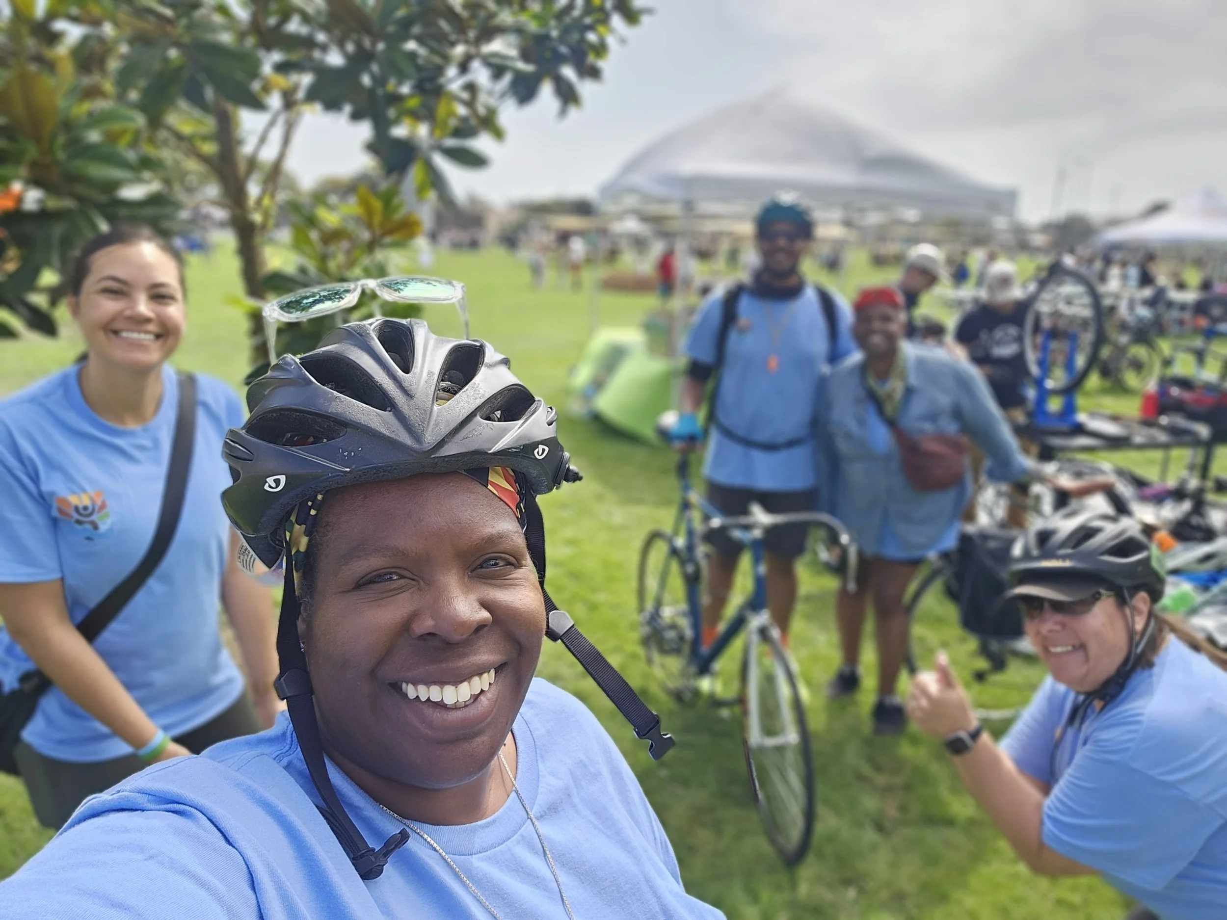 A group of smiling people in bike helmets and casual clothes gathered outdoors at a biking event, with bikes in the background.