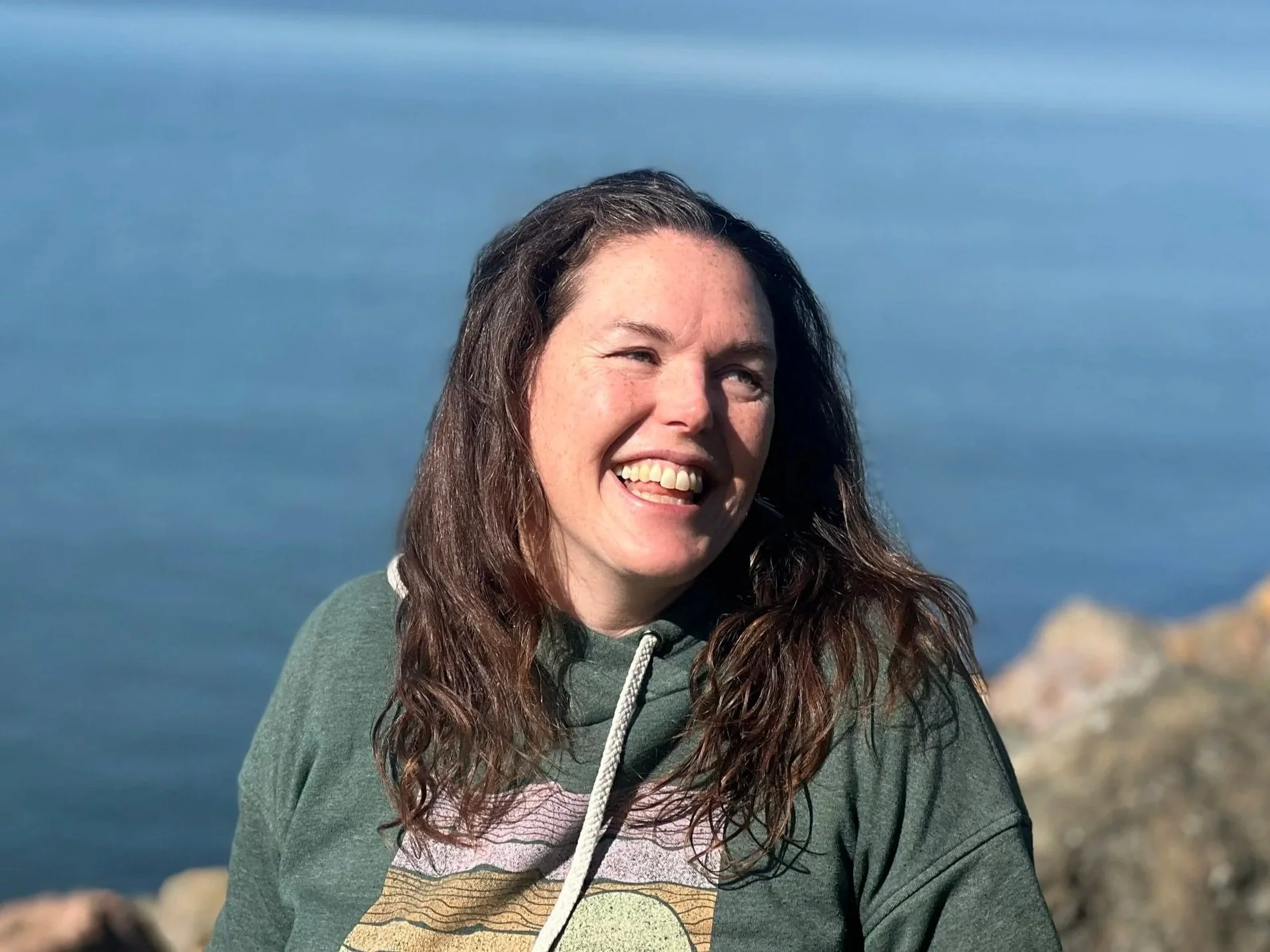 Smiling woman wearing a green hoodie sweatshirt, in the sun, in front of rocks and water. 
