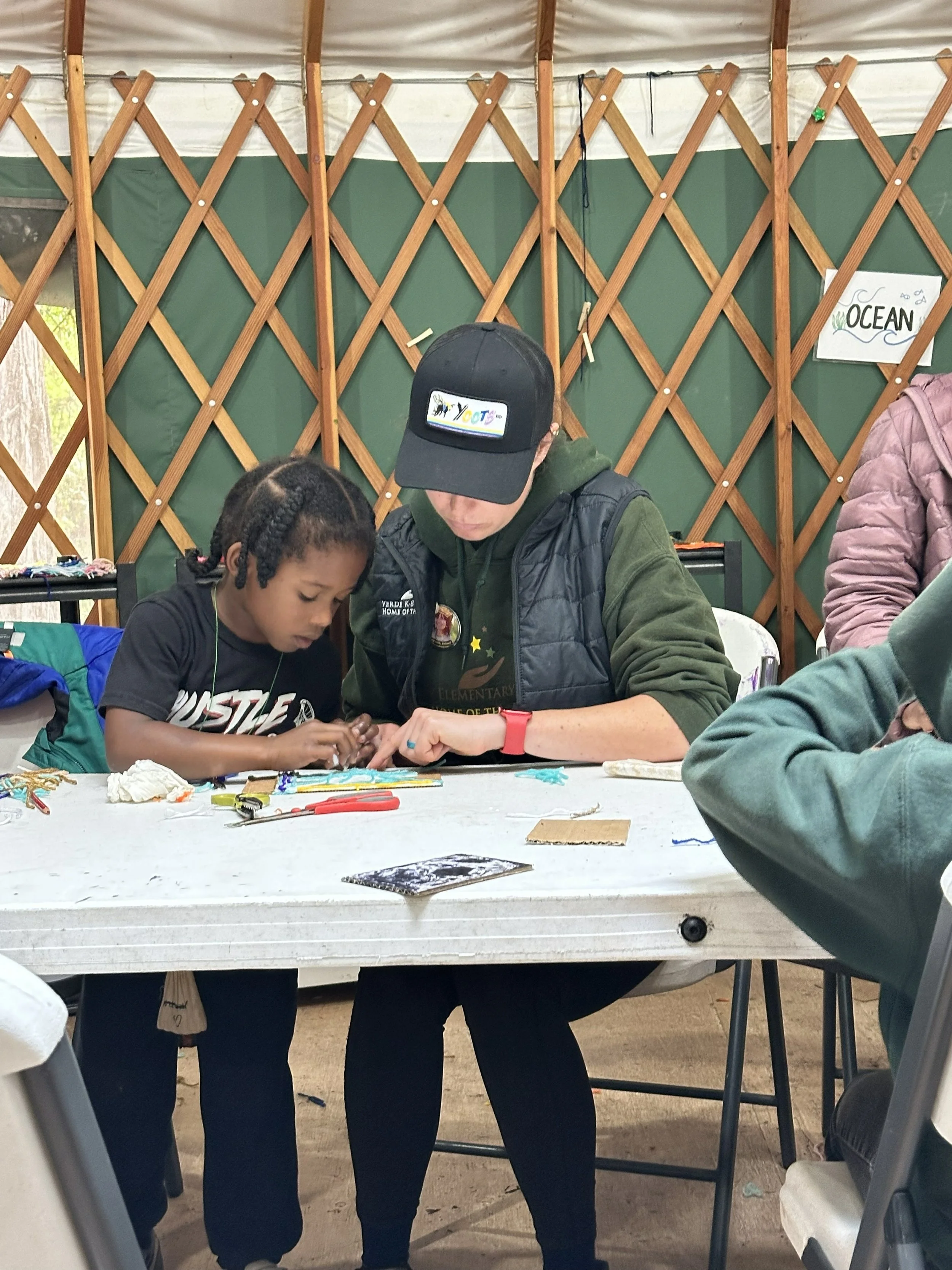 A young boy and an adult sit at a table working on a craft project inside a yurt with wooden lattice walls. They are focused on their work, with craft supplies spread on the table. 