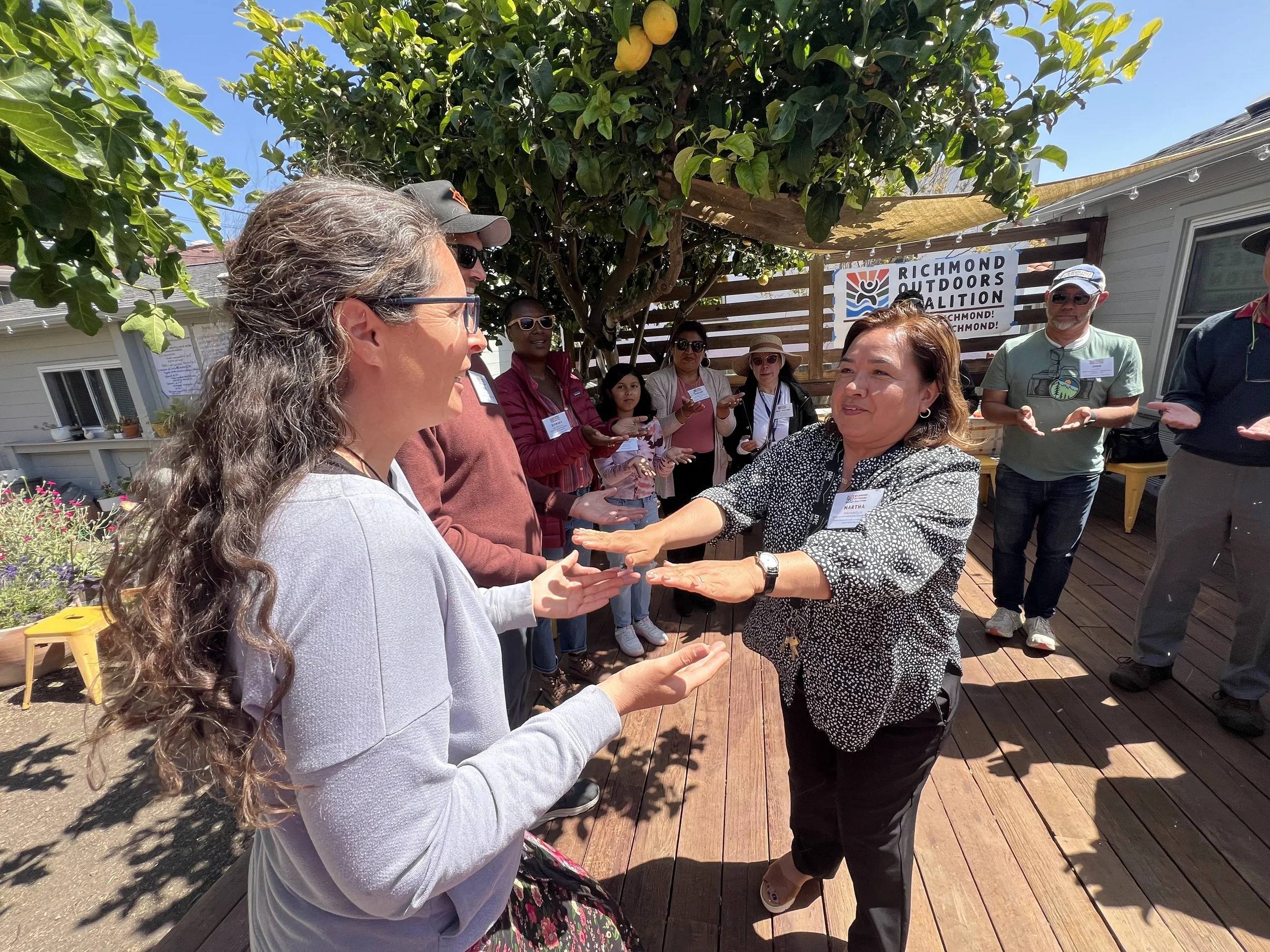 A group of people are gathered outdoors on a wooden deck, under a large tree with green leaves and yellow fruit. Two women in the foreground are playing a game with hand gestures.