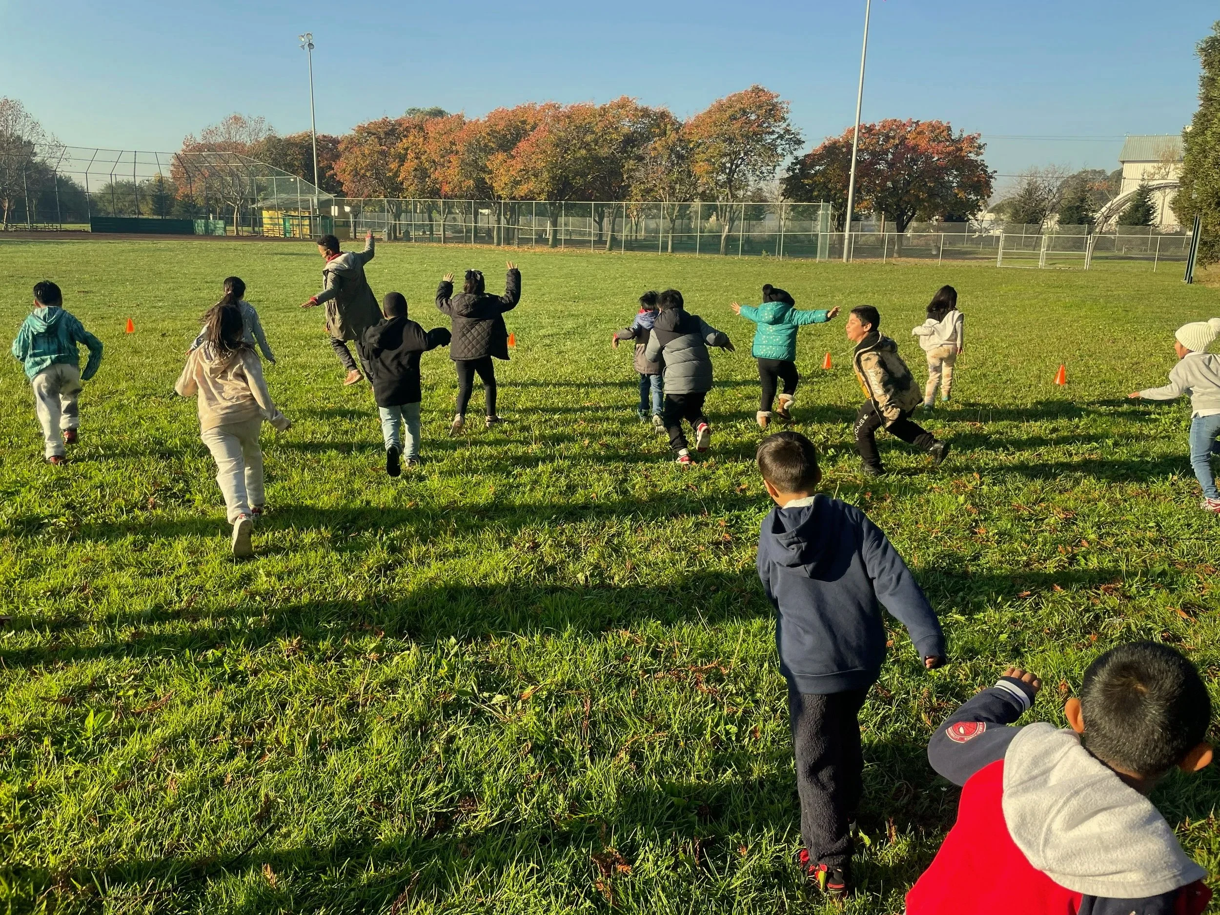 Group of children running in a field with a woman, with their arms out, with trees and a metal fence in the background.