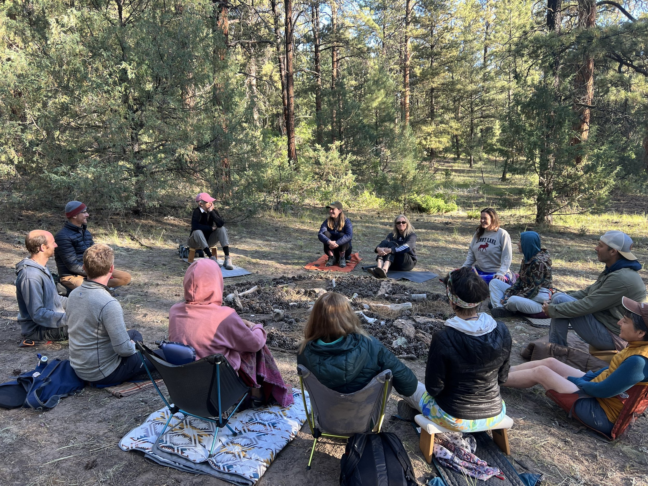 13 people sitting outside in a circle on meditation cushions, blankets and chairs, looking at one person who appears to be teaching, with a nature mandala made of pine needles, stones and sticks in the center of the circle. 