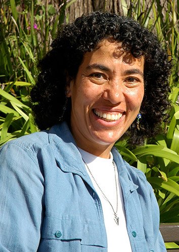 A smiling woman in the sunshine, wearing a white shirt underneath a blue collared shirt, in front of green leaves.