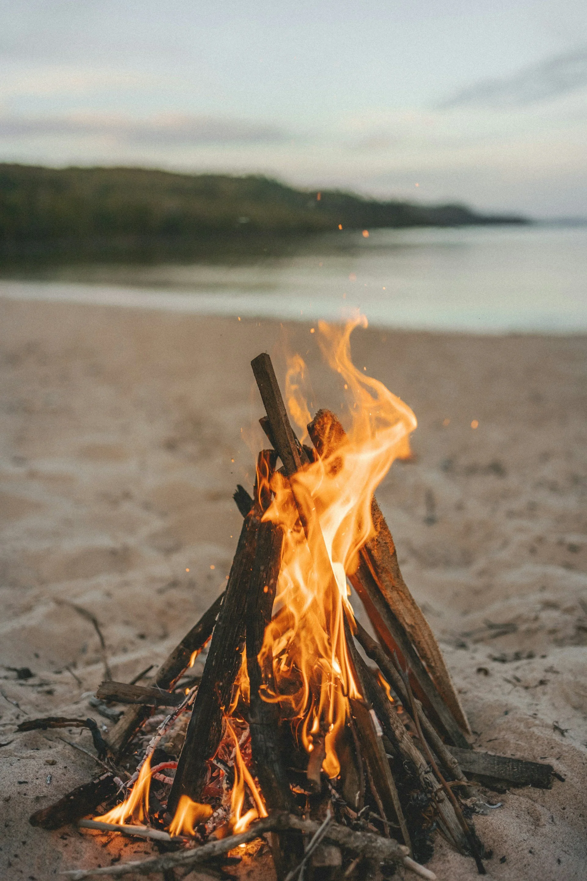 Campfire burning on a sandy beach with water in the background.