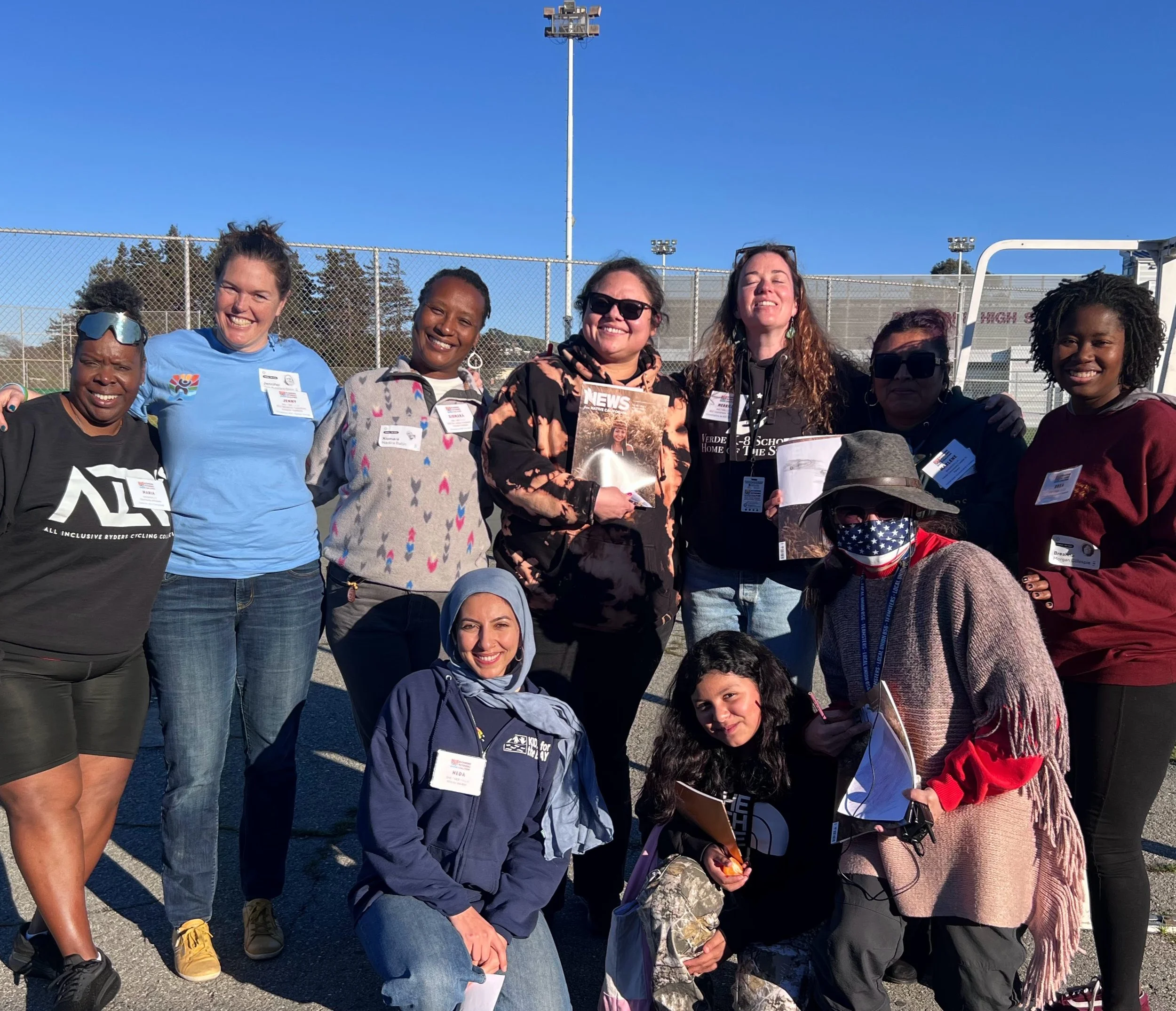 10 people standing and squatting with their arms around each other, smiling and looking at the camera, on a sunny day with a sports field and blue sky in the background. 