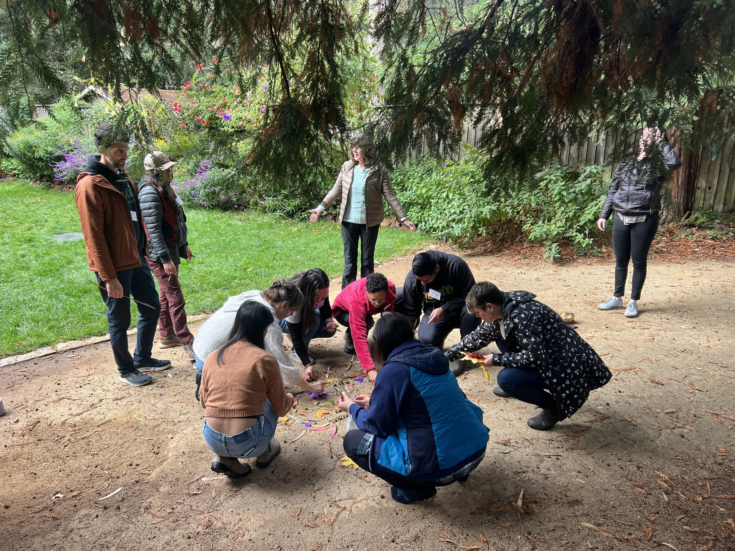 Group of adults outside, crouched in a circle, laying out natural materials on dirt, with grass, flowers and trees in the background and a few adults standing nearby watching.