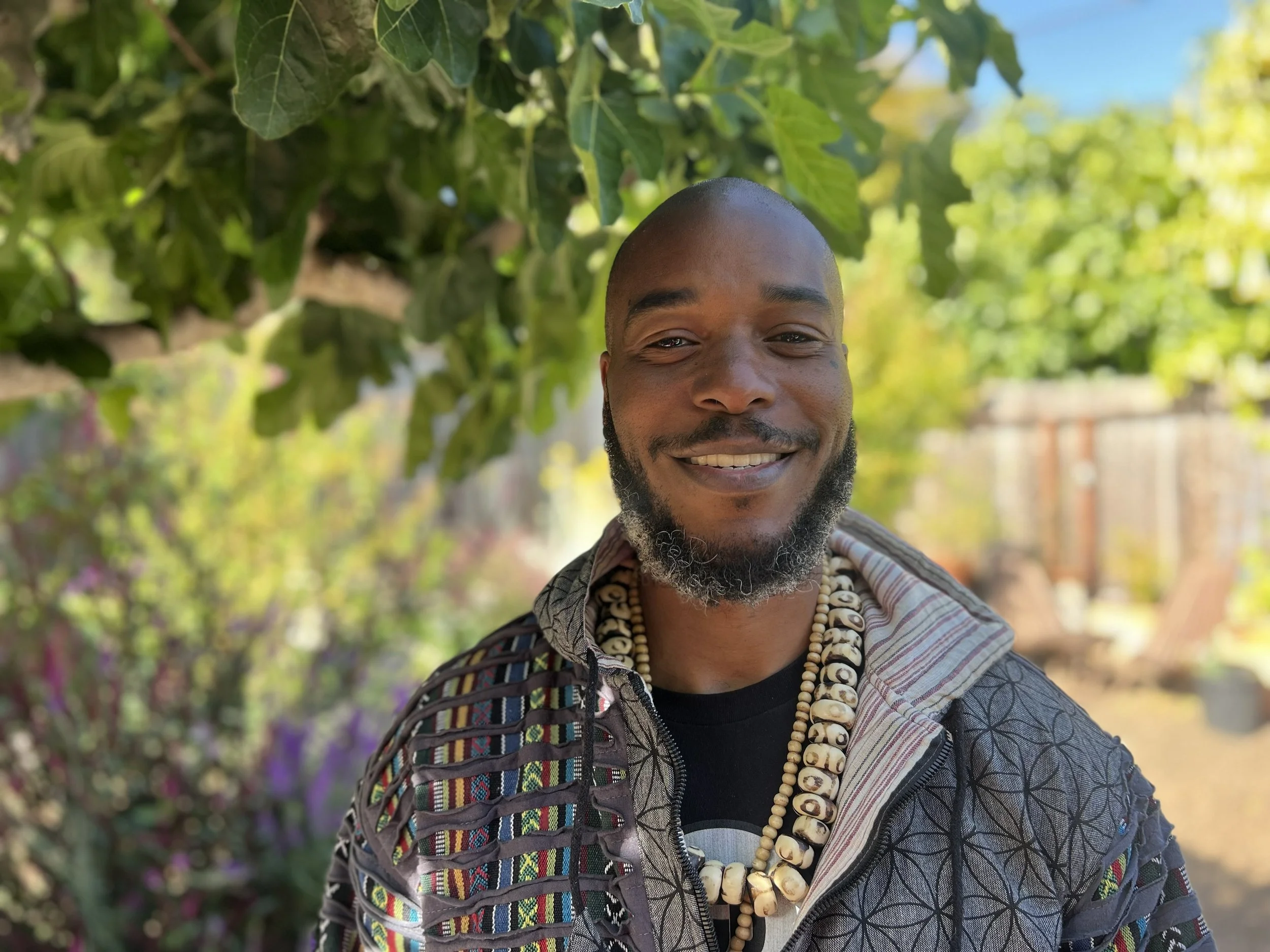 Smiling man wearing a black t-shirt, shell necklaces and a multicolored jacket, in front of green leaves and a blurred green background. 