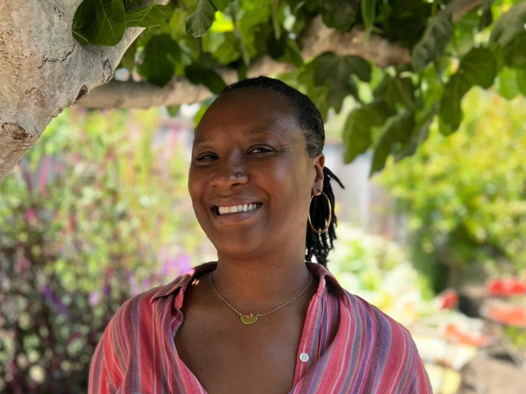 Smiling woman wearing a pink button down shirt, in front of green leaves and a blurred green background. 