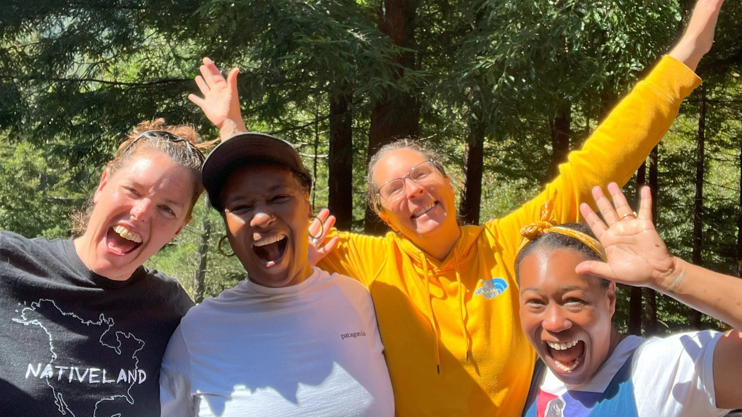 Four women are smiling and having fun outdoors in a green forest. They are close together, with one woman in a black t-shirt, one in a white t-shirt and cap, one in a yellow hoodie, and one in a white and blue top. They are making cheerful gestures with their hands.