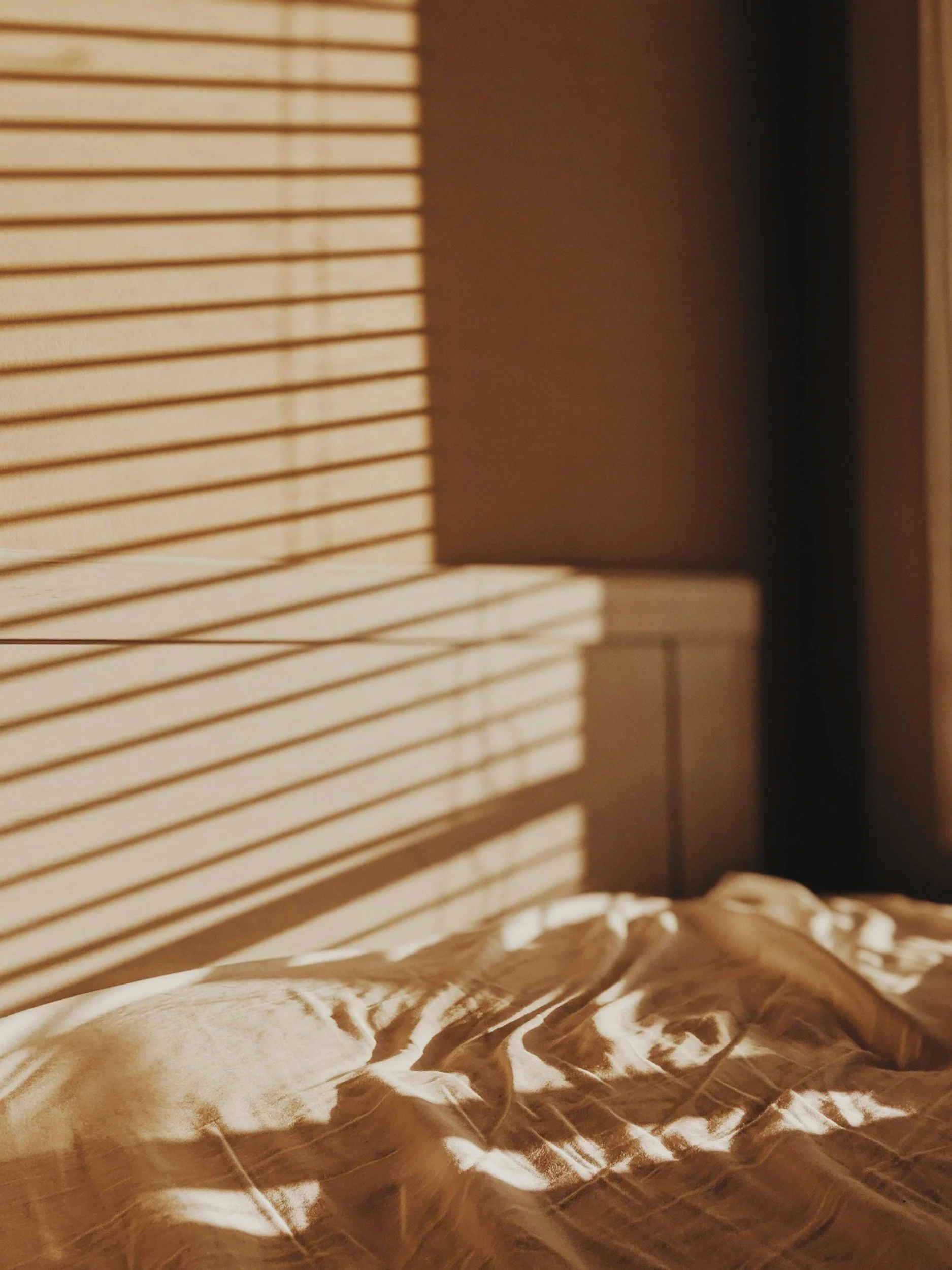 Sunlight streams through window blinds creating striped shadows on a bed with beige crumpled bedding in a warmly lit room.