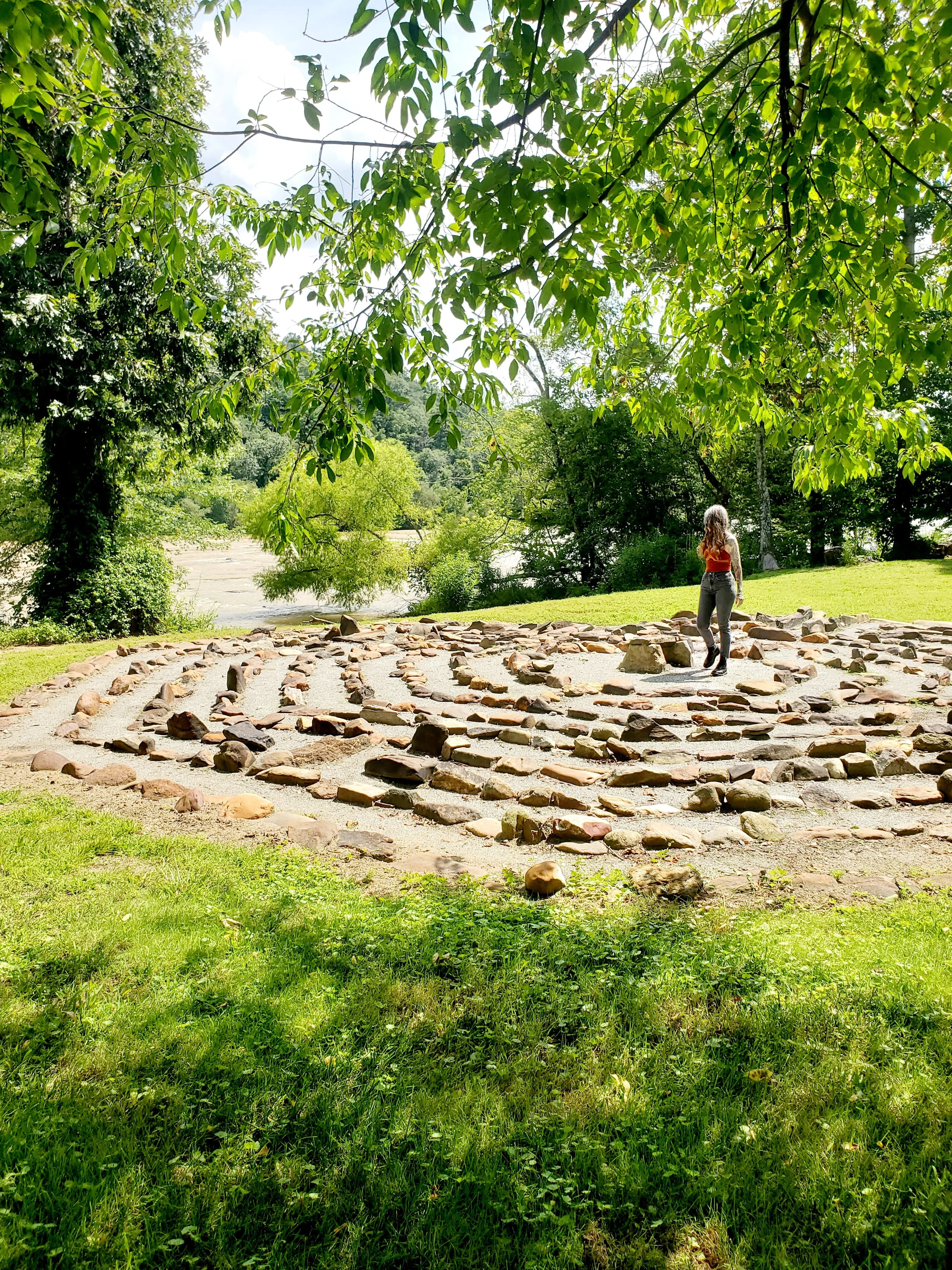 A person with long curly hair, wearing a red top and gray pants, walking on a circular rock arrangement in a lush green park with trees and grass.