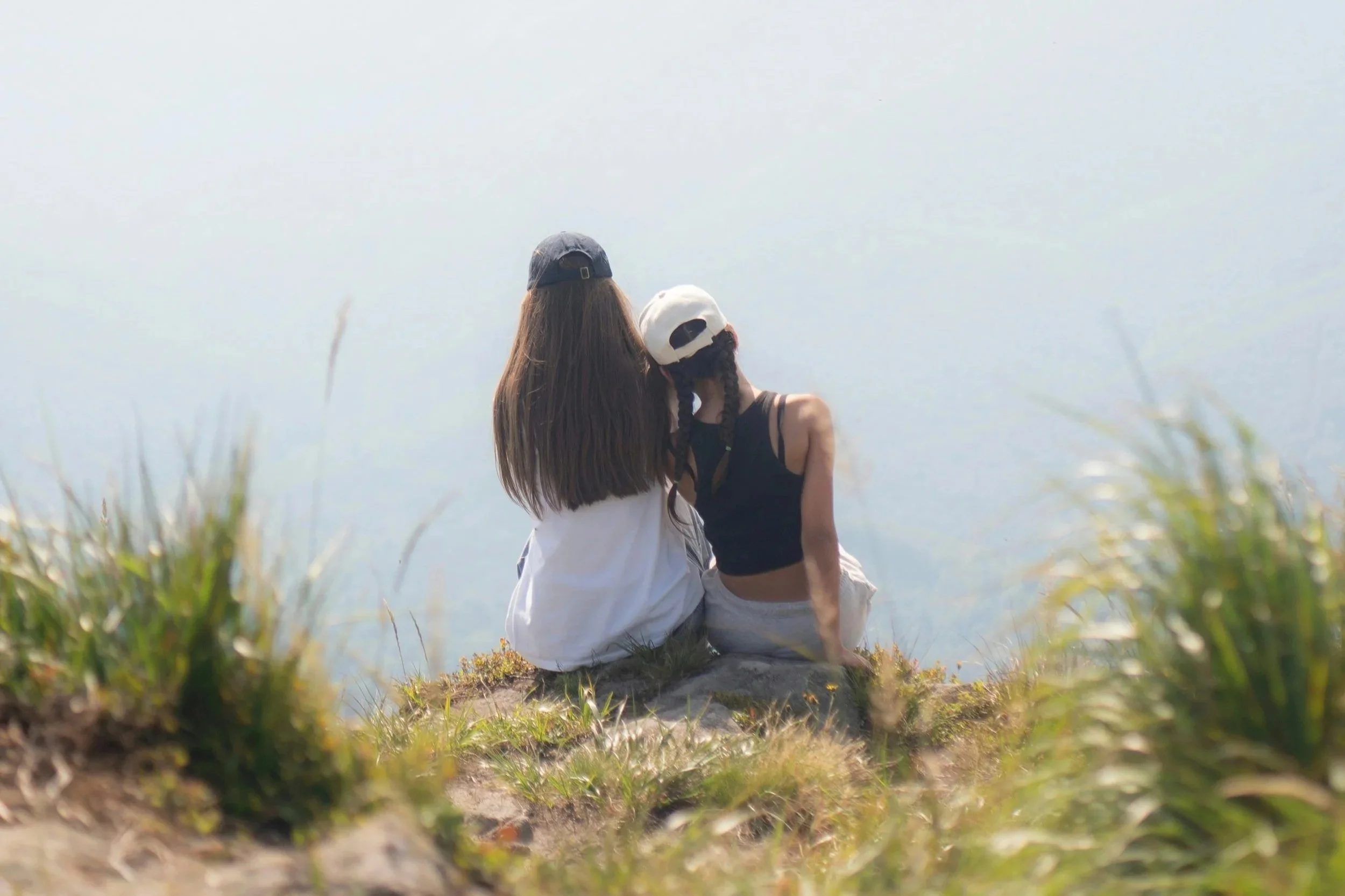 Two girls sitting on a scenic overlook with their backs to the camera. One has her head on the other's shoulder.