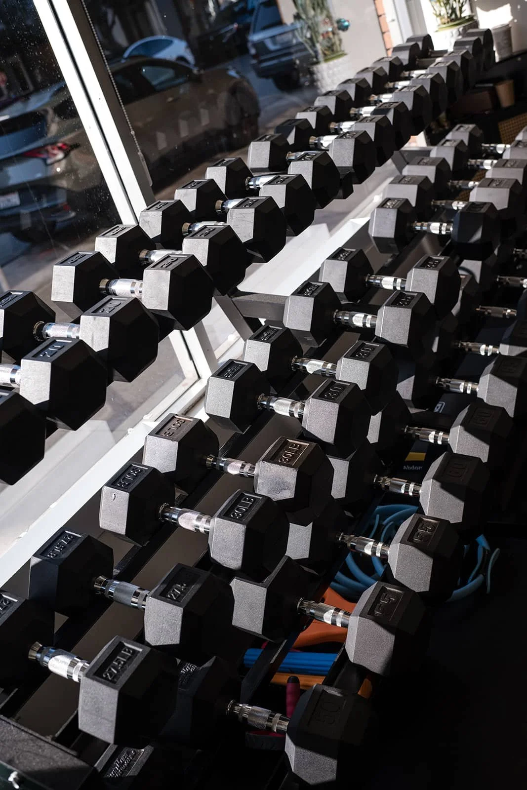A row of black and gray dumbbells on a black rack inside a gym, with large windows showing parked cars outside.