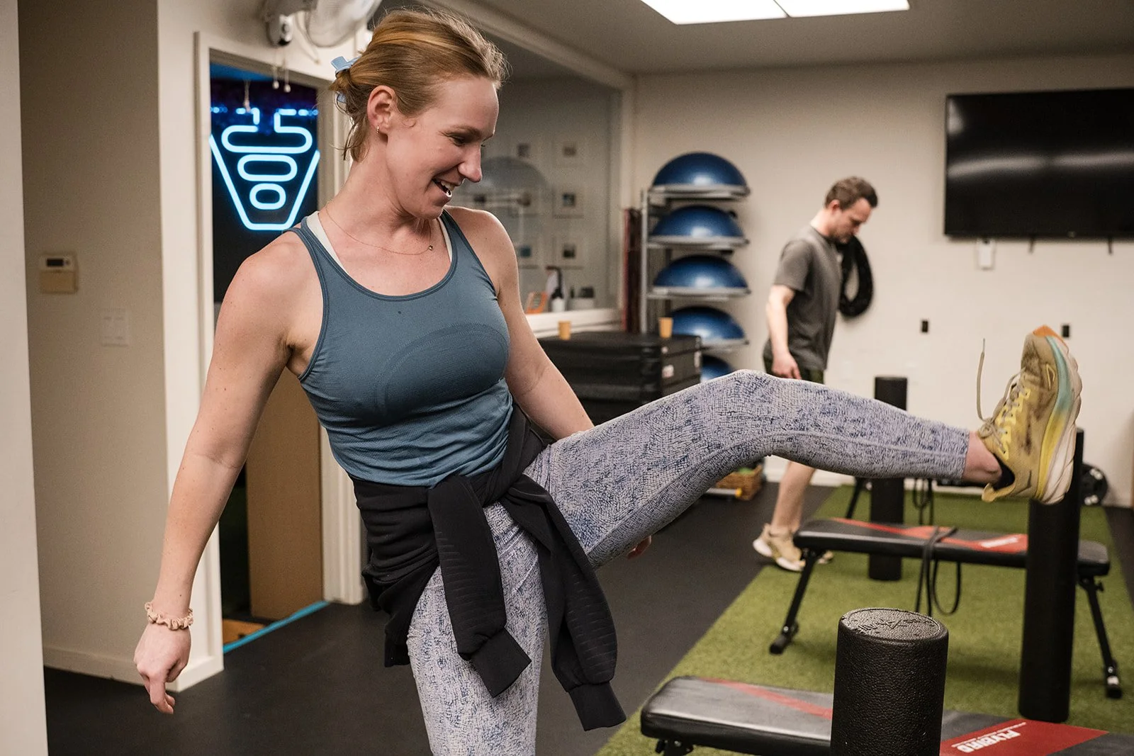 A woman with a smile wearing athletic clothing and yellow sneakers kicks her leg out as part of a small group fitness class.