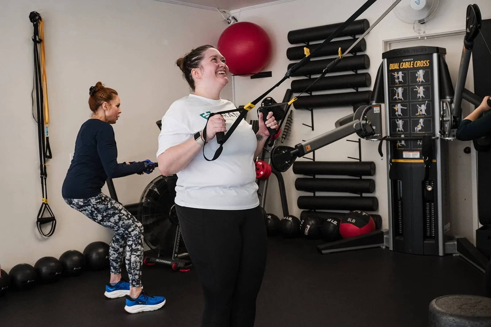 Two women working out in a gym. The woman in front is smiling, holding TRX suspension straps. The woman in back is squatting, using a kettlebell. Gym equipment, including medicine balls, foam rollers, and resistance bands, is visible.