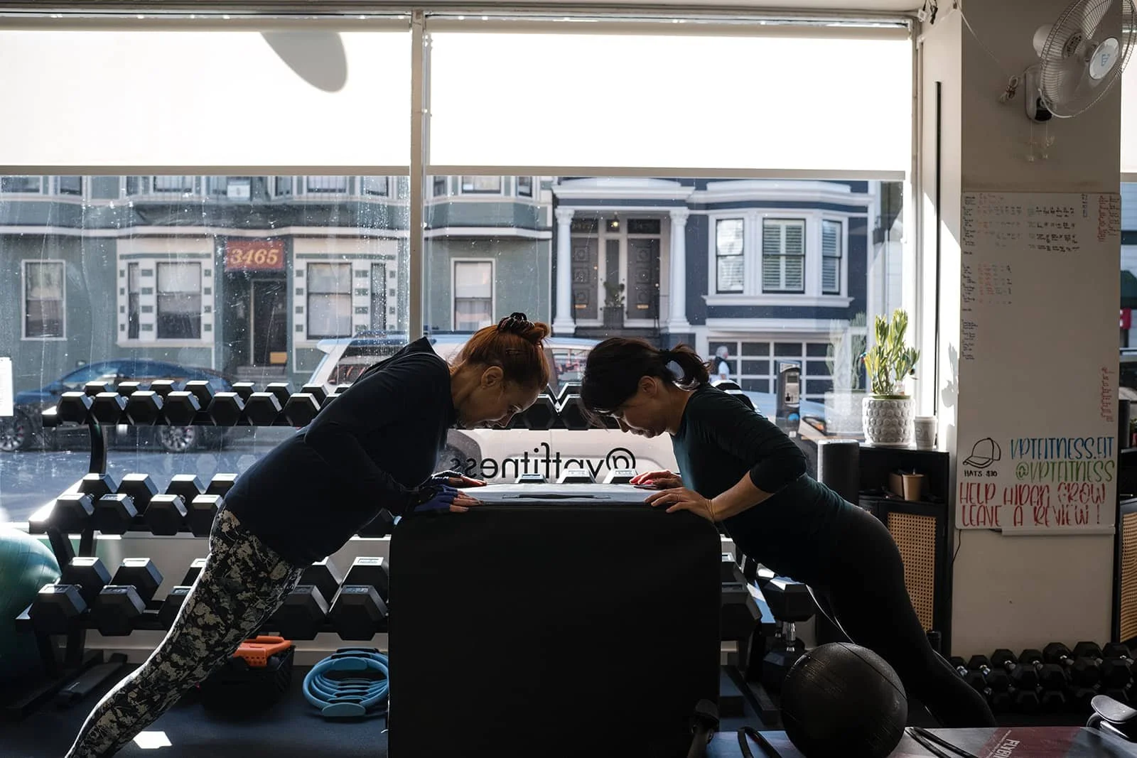 Two women in workout attire perform a plank exercise in a fitness studio with lots of natural light coming in.