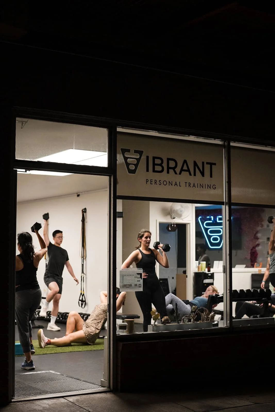 People exercising at a personal training gym, viewed through a large front window, with a sign that reads 'VIBRANT Personal Training' outside.