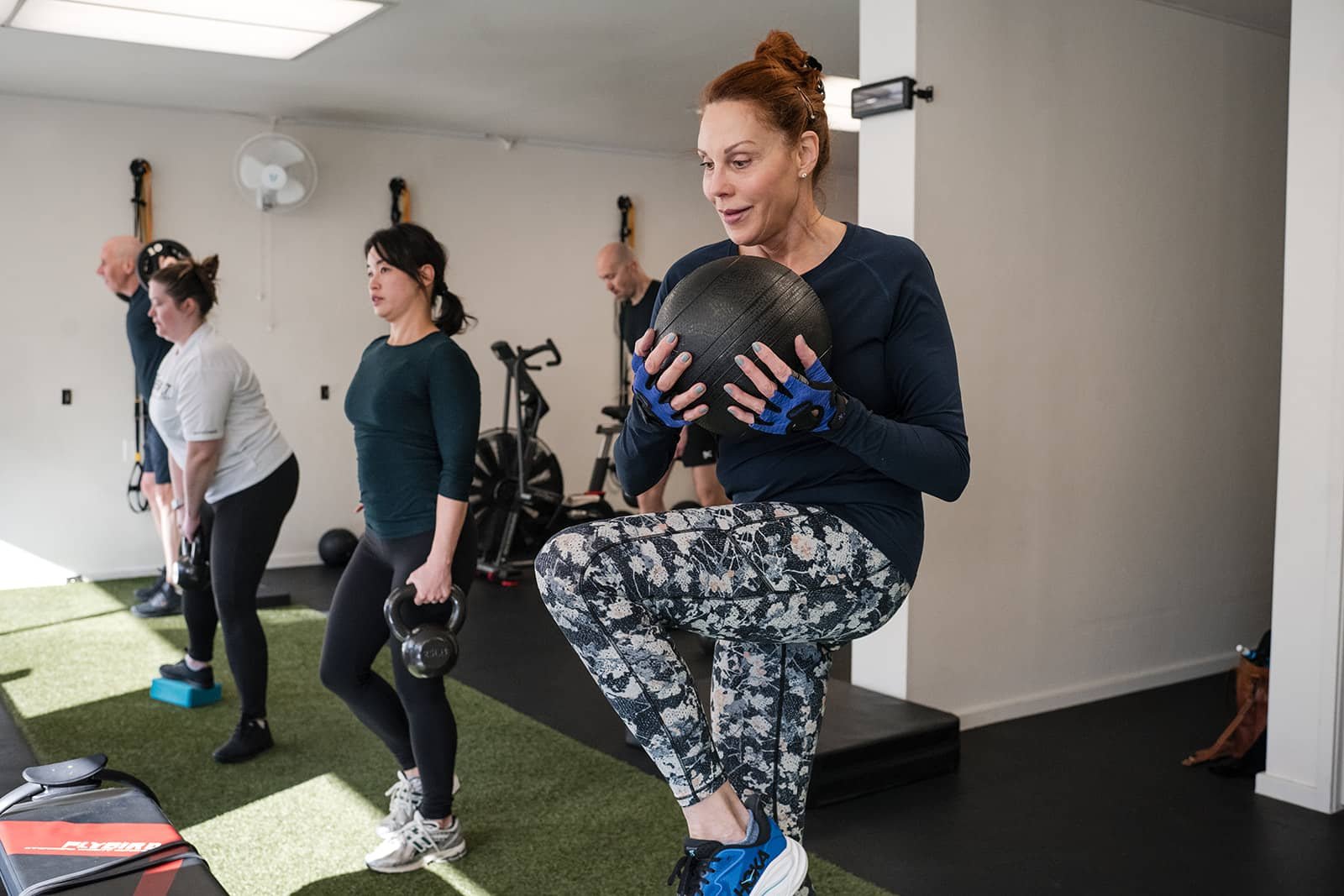 A woman holding a black medicine ball while balancing on one leg during a workout session in a gym with other people exercising in the background.