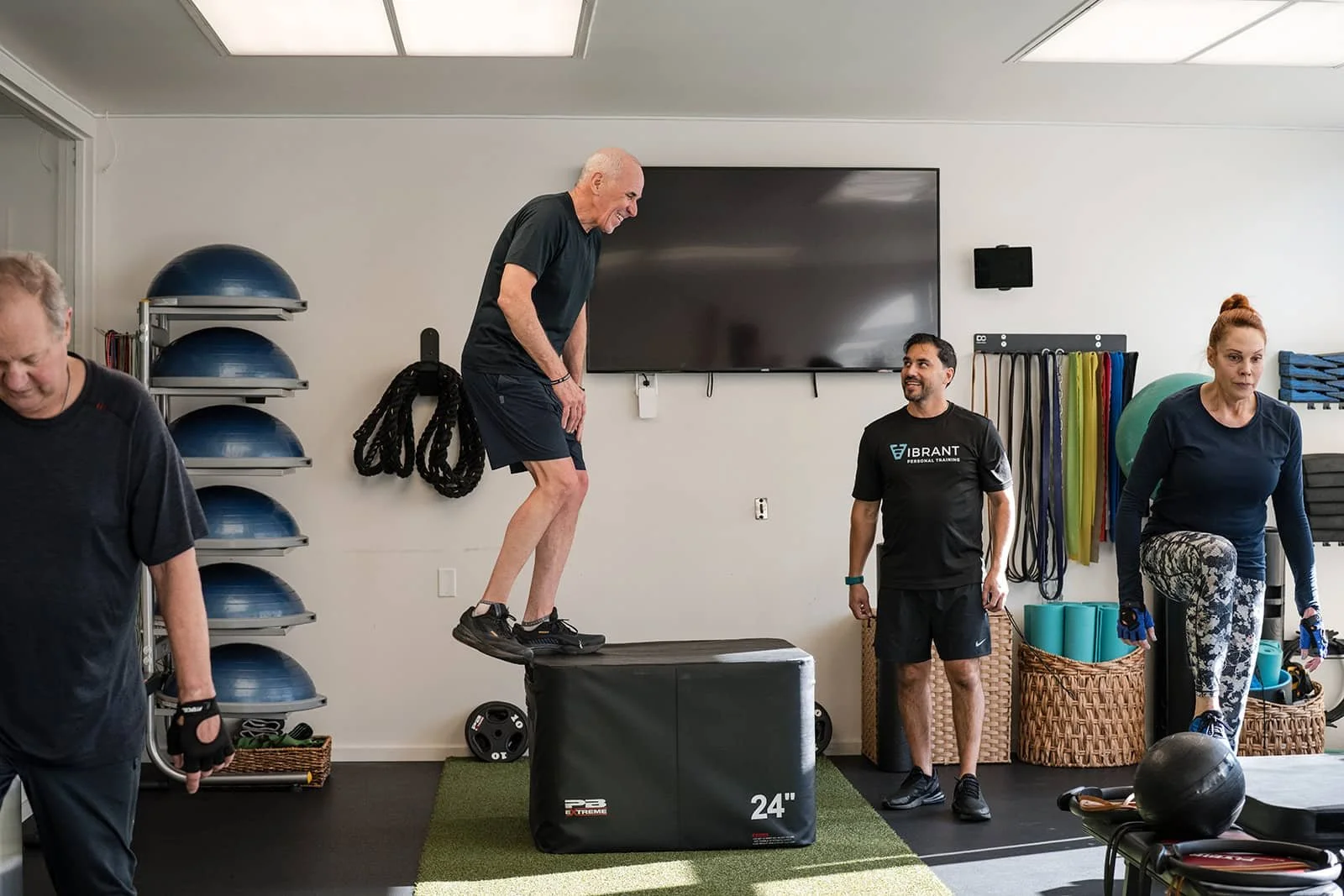 People exercising in a gym, one man jumping onto a box, others standing nearby with various workout equipment in the background.