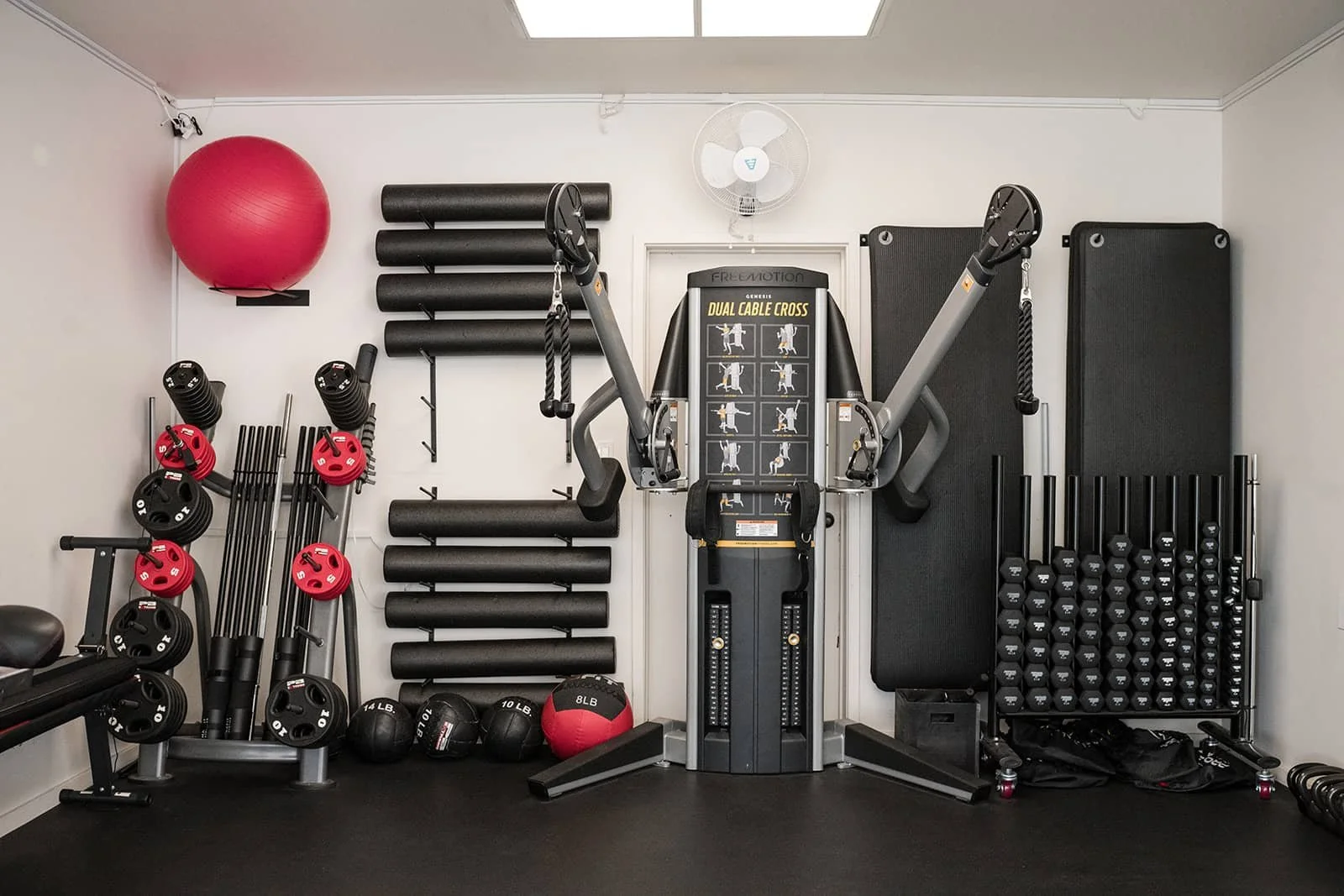 An indoor gym with various fitness equipment, including red and black weight plates, medicine balls, foam rollers, and a dual cable cross workout machine, with black wedges on the back wall and a wall-mounted fan.