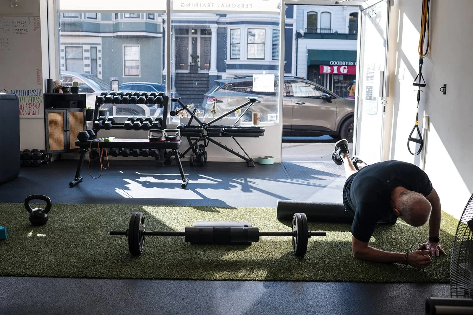 An indoor gym with a man performing a plank on a green mat, a barbell on the floor, a kettlebell and a small plank on the side. The gym has a large front window with sunlight, a bench with dumbbells, and various workout equipment. Outside, a parked c