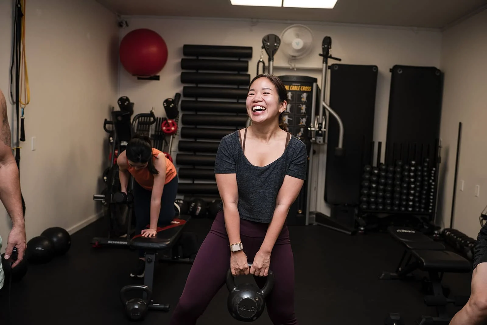 A woman smiling and holding a kettlebell inside the fitness studio, with training equipment and another person working out in the background.