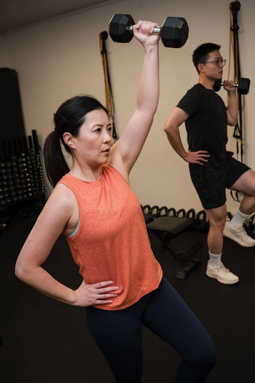 A woman in an orange workout tank top and black leggings lifting a dumbbell overhead in a gym, with a man in the background performing a similar exercise.