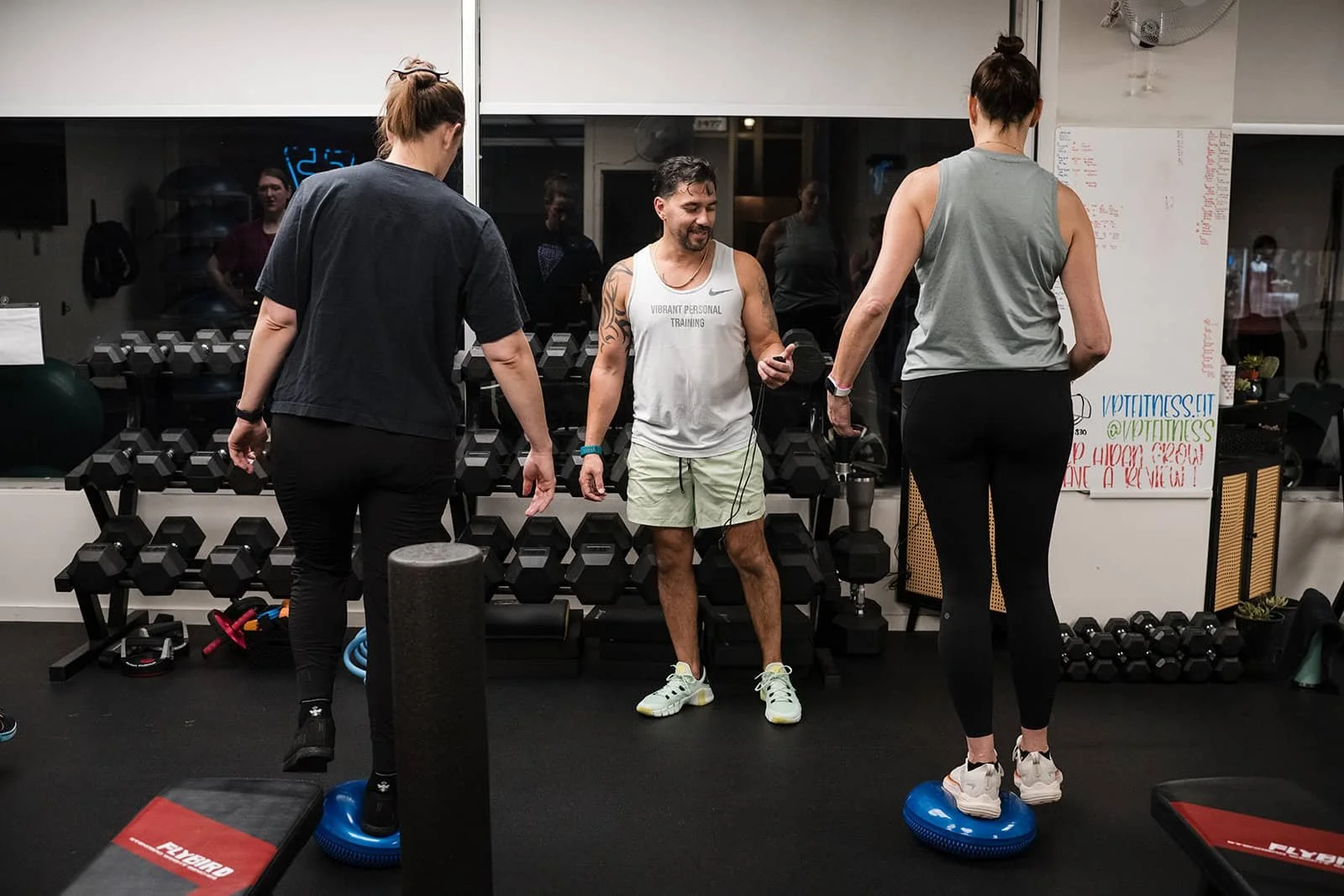 Two women standing on balance disks while the trainer instructs. Gym equipment, weights, and a whiteboard are visible in the background.