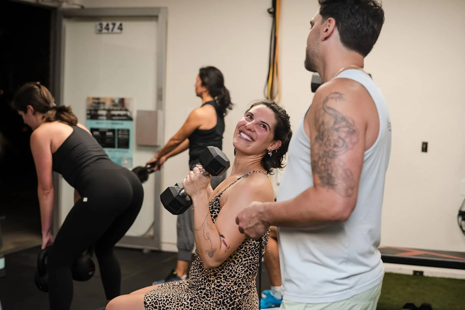 A woman smiling while lifting a dumbbell in a gym, surrounded by other people exercising.
