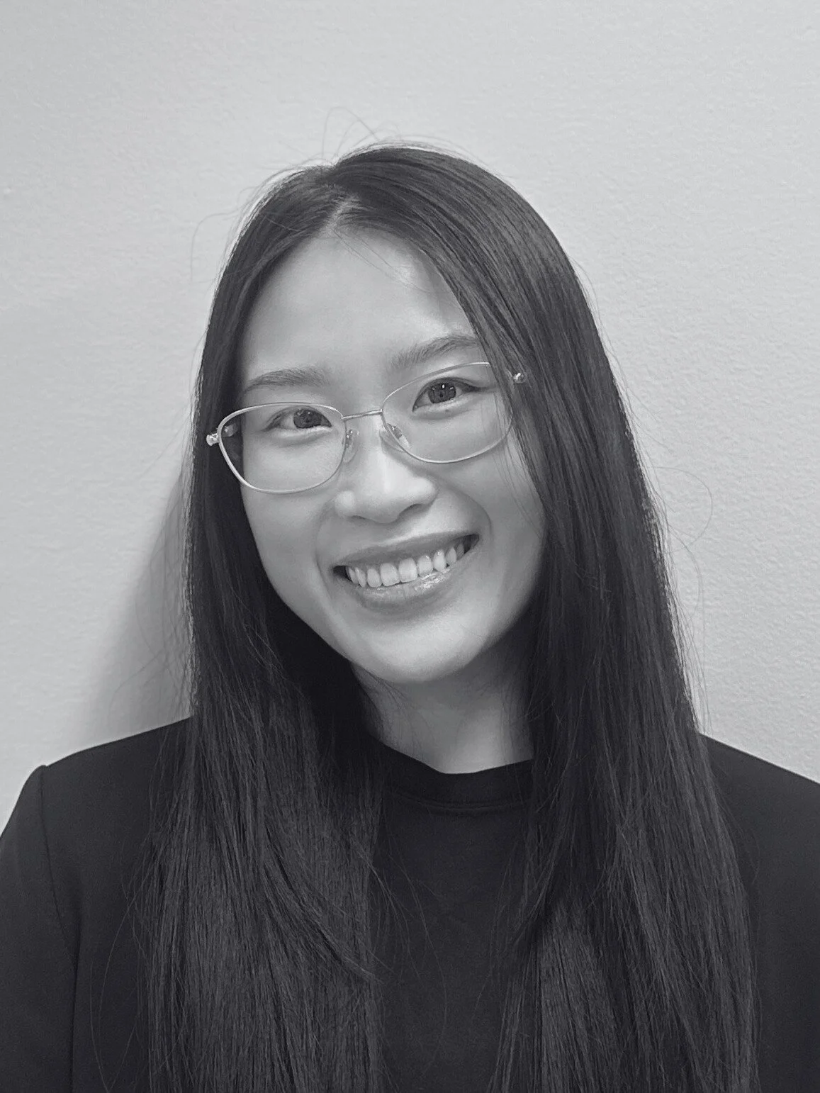 Black and white photo of a smiling woman with long straight hair, wearing glasses and a dark top, standing against a plain wall.