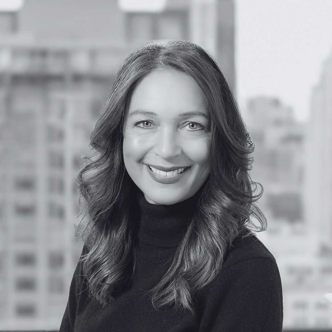 Black and white portrait of a smiling woman with long wavy hair, wearing a dark turtleneck sweater, with a cityscape background.