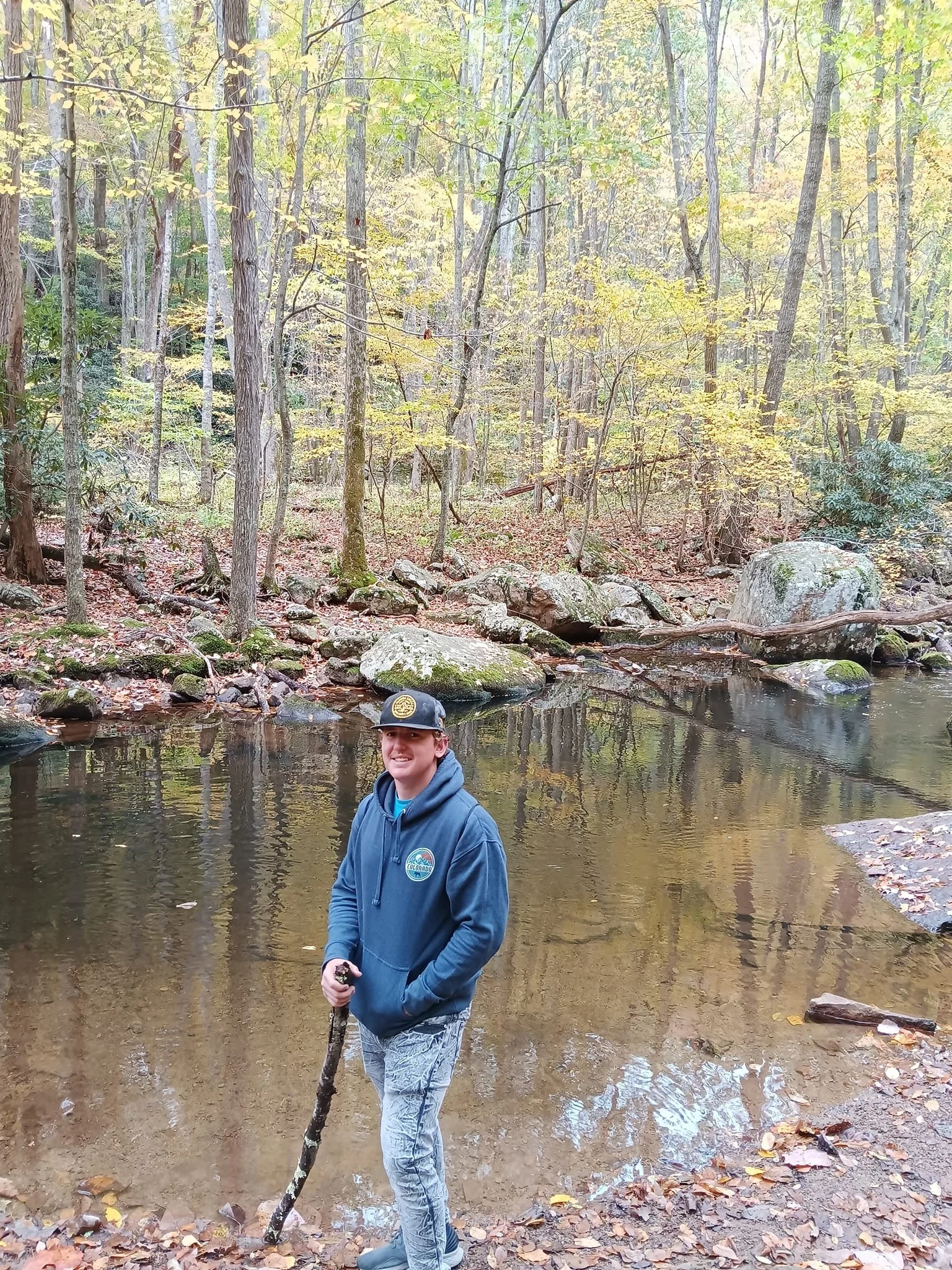 A young man standing by a creek in a forest, holding a stick, wearing a hat and a blue hoodie.