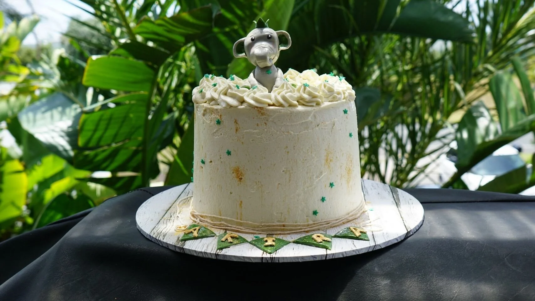 White birthday cake with green star sprinkles and a monkey cake topper, sitting on a decorated cake board with green and gold letters, against a background of green plants.