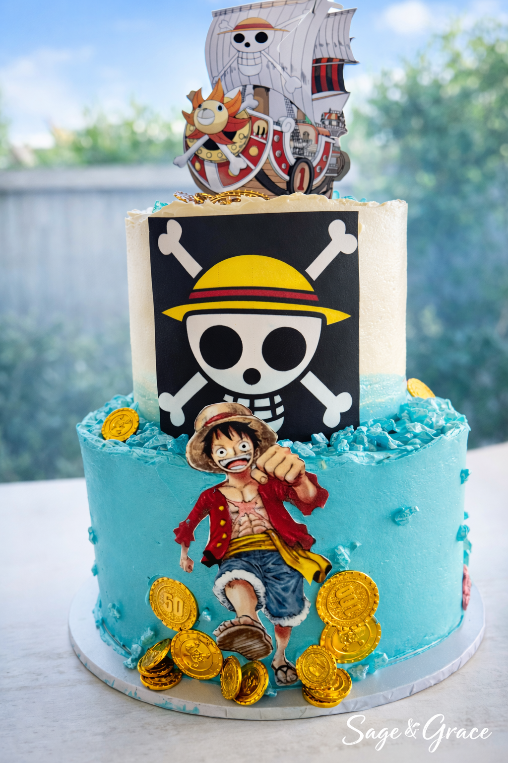Two-tiered birthday cake with One Piece theme, featuring character decorations including Luffy and the Straw Hat Pirates logo, gold coin toppers, and an ocean blue frosting.