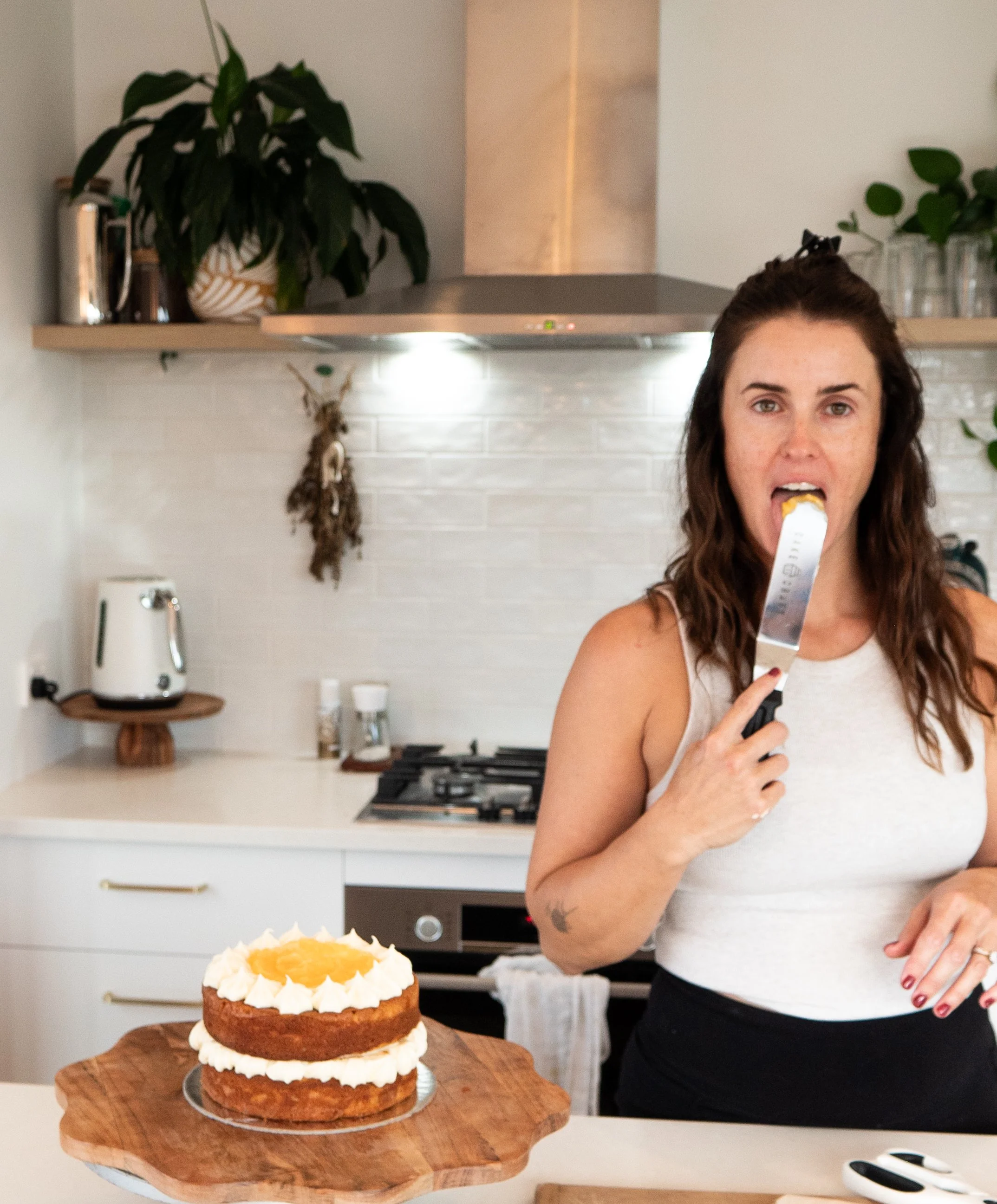 Woman in a white tank top holding a cake knife in her mouth in a modern kitchen with a layered cake on a wooden serving board in front of her.