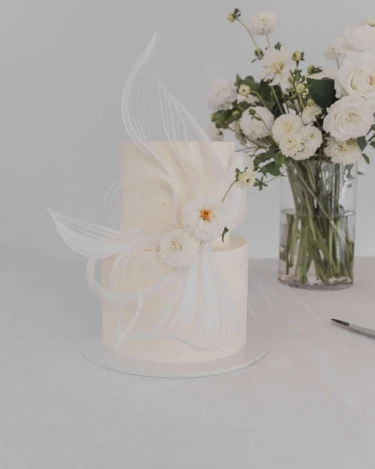 A white cake decorated with delicate white flowers and white, feather-like sugar art, placed next to a vase of white and pink flowers.