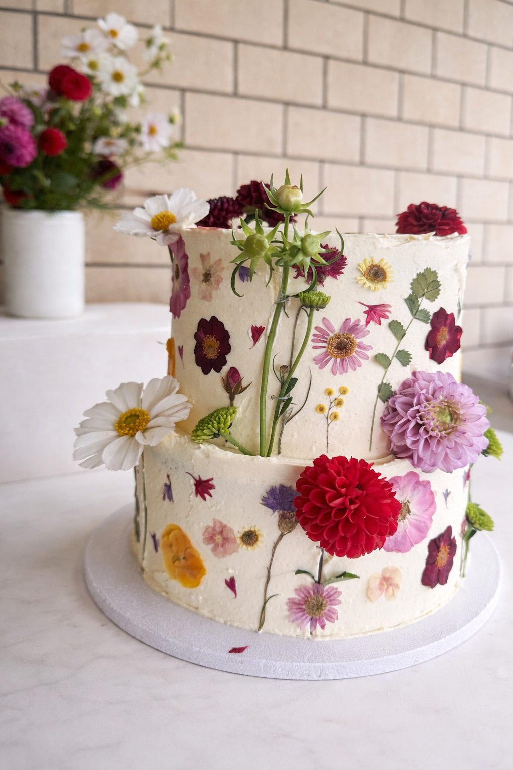 A two-tiered cake decorated with real and edible flowers against a white table and brick wall background.
