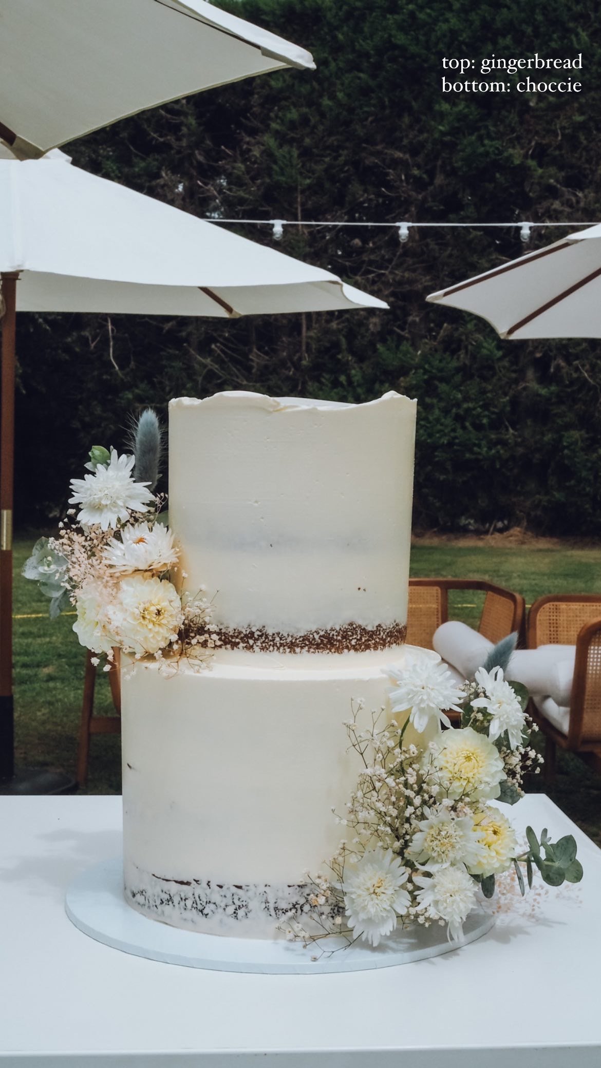 A two-tier white wedding cake decorated with white flowers and greenery, set outdoors under umbrellas.