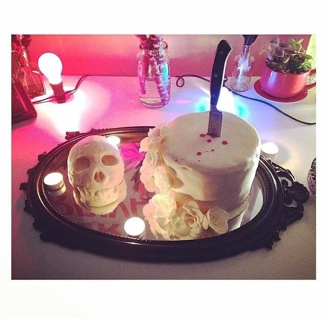 A white skull-shaped candle and a white heart-shaped cake with a knife on top, decorated with white roses and pink dots, are placed on a mirror tray surrounded by small round lights on a vanity table.