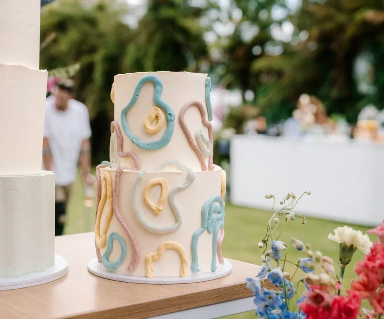 Two-tiered white cake decorated with pastel-colored abstract icing designs, set outdoors at a celebration or event, with blurred flowers and other tables in background.