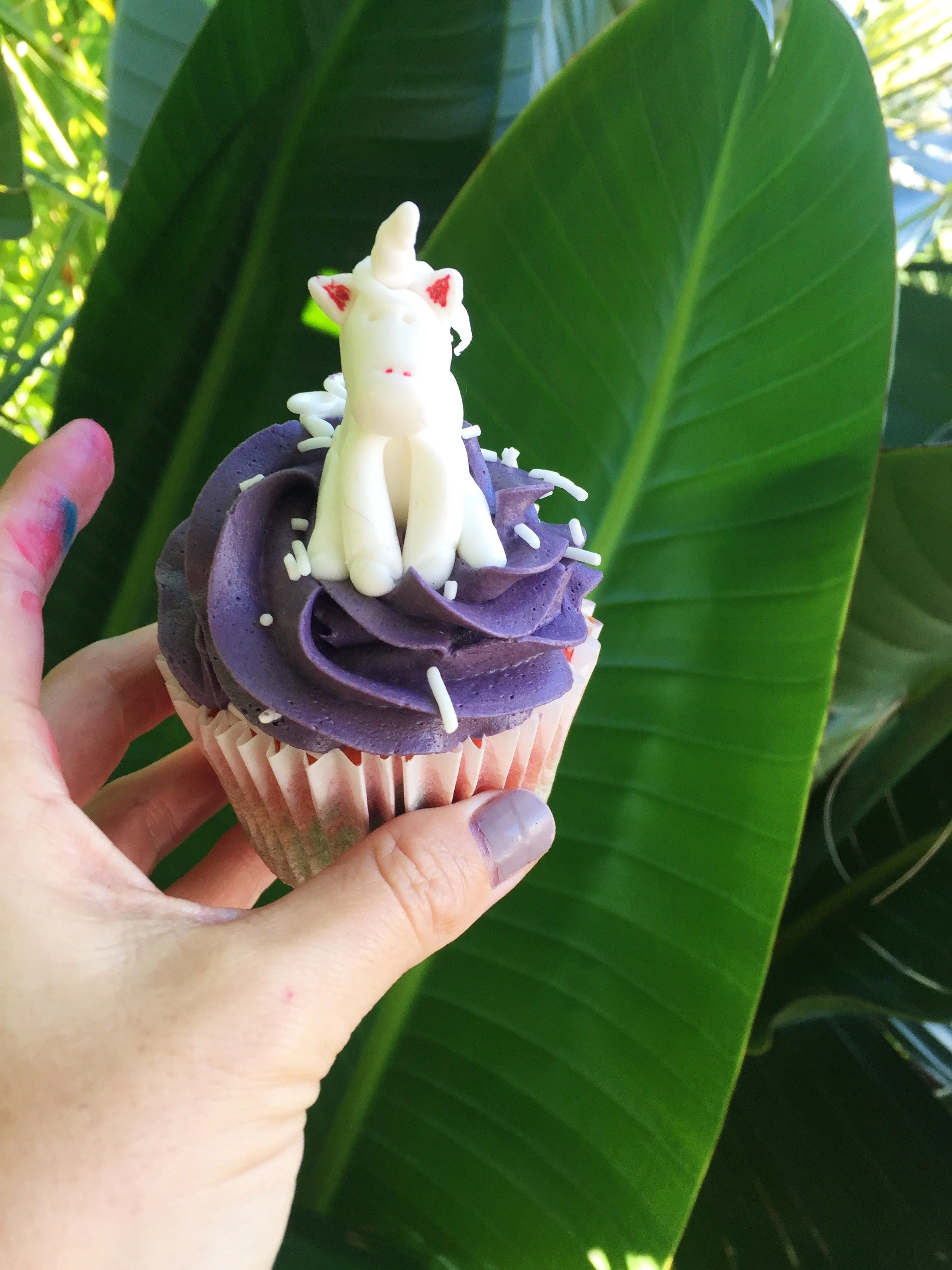 Hand holding a purple frosting cupcake topped with a white unicorn figurine, sprinkles, and a large green leaf in the background.