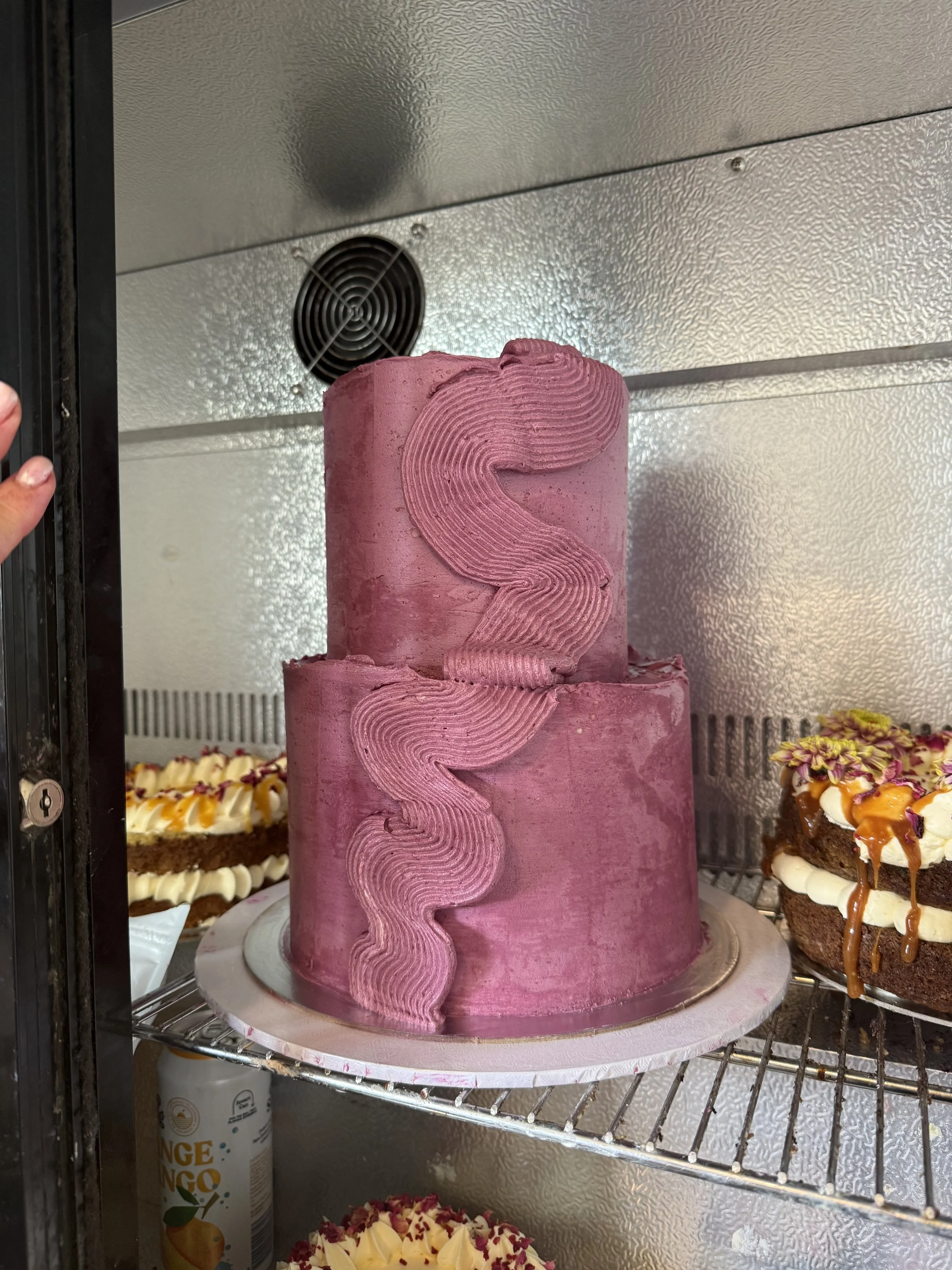 Pink two-tiered birthday cake with decorative icing on a cake stand in a display case.
