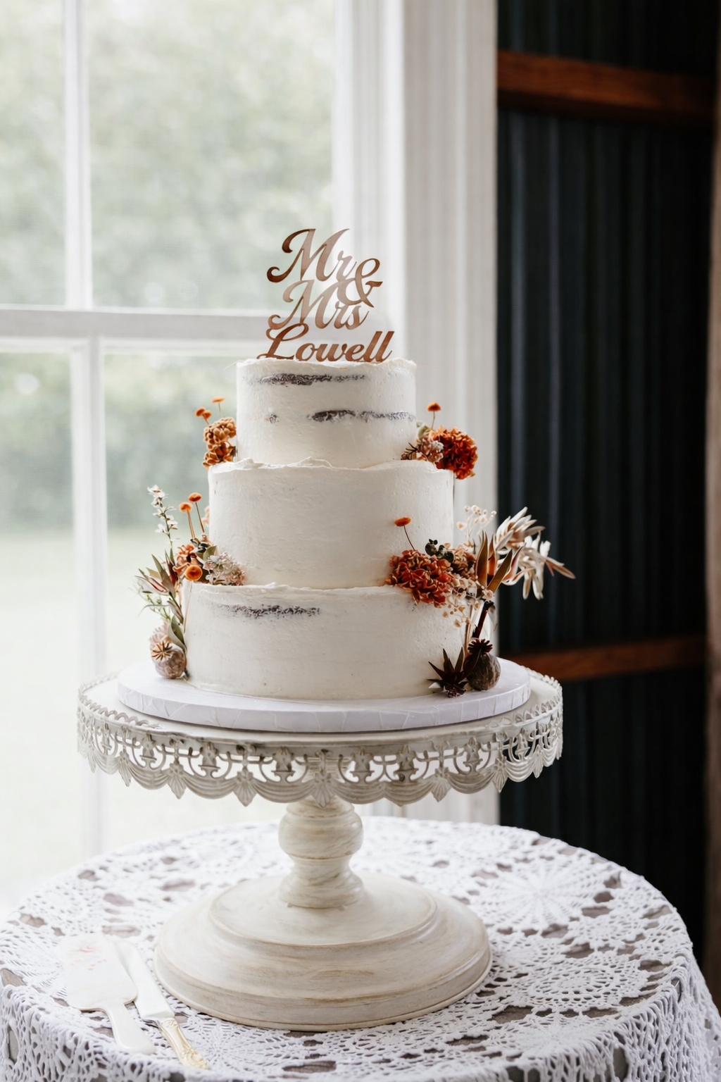 A three-tiered wedding cake with white frosting, decorated with orange and white flowers, and a wooden topper reading 'Mr & Mrs Lowell' on a vintage cake stand with lace tablecloth.