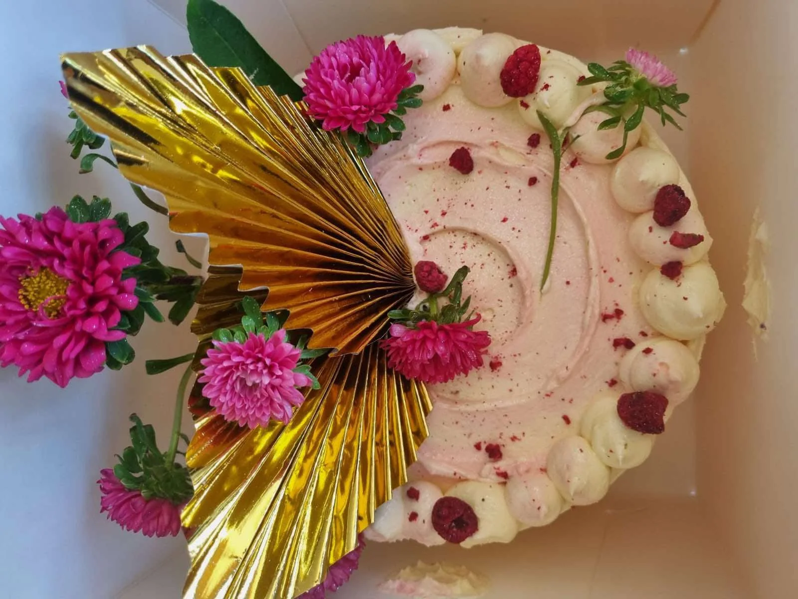 A decorated cake with pink and white frosting, raspberries, and edible flowers, adorned with gold foil decorations.