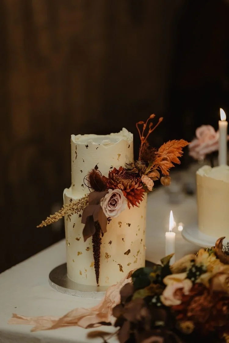 A two-tiered white wedding cake decorated with dried flowers and gold leaf on a table, with candles and floral arrangements nearby.