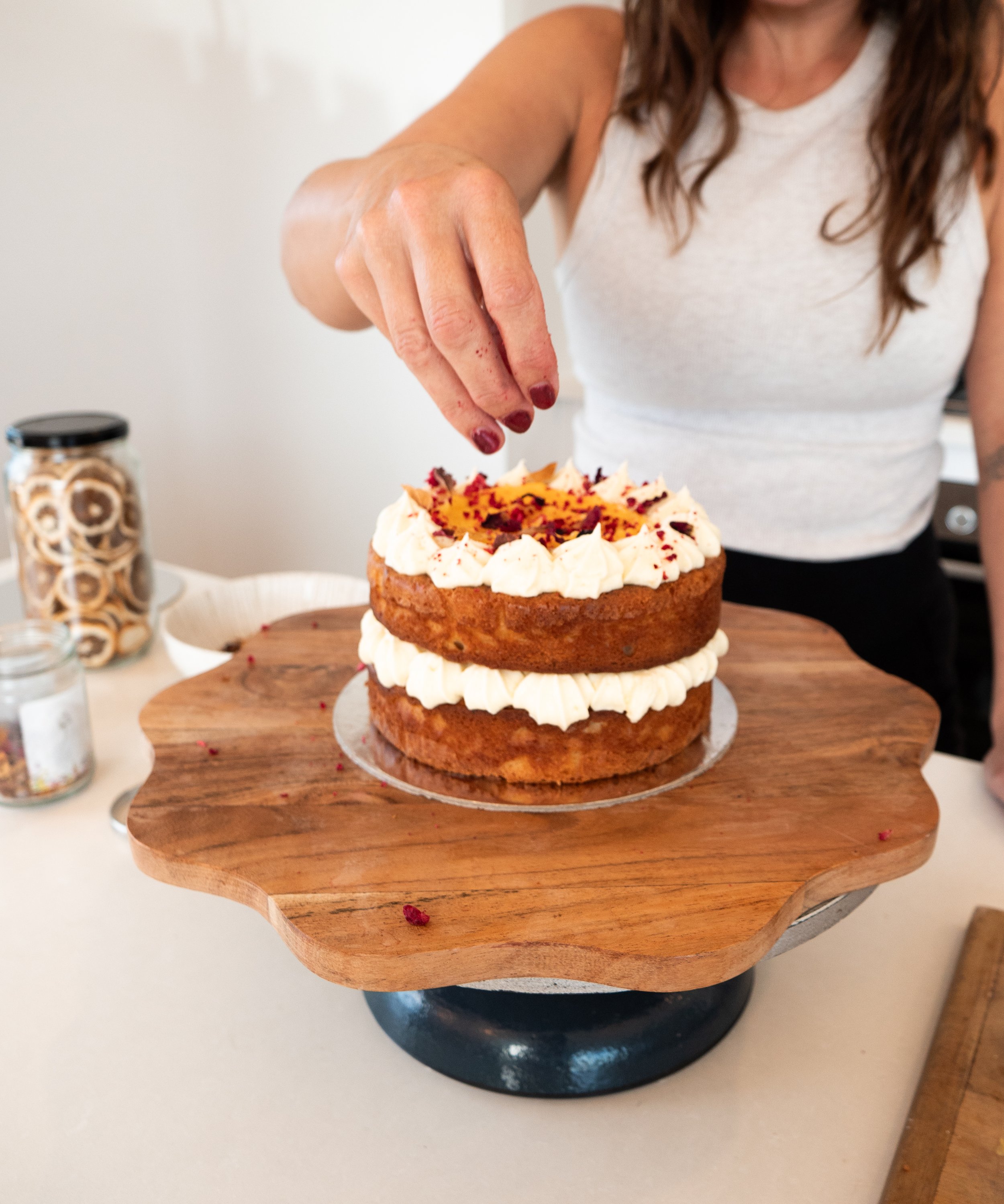 Woman decorating a two-layer cake with white frosting and topping with dried fruits.