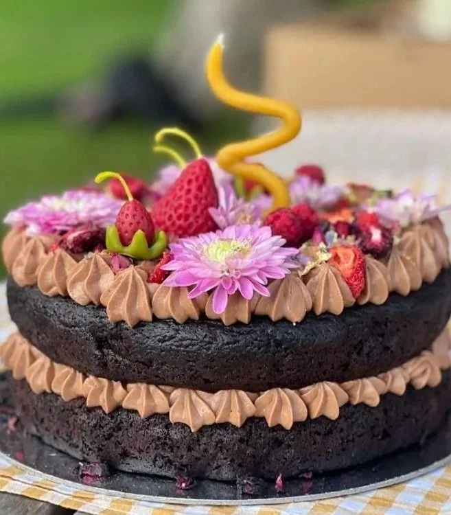 A two-layer chocolate cake decorated with pink flowers, strawberries, and a yellow spiral candle.