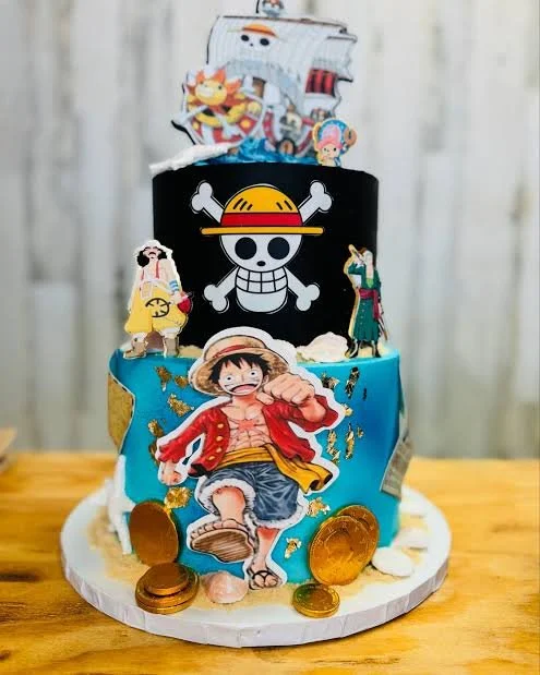 A colorful cake decorated with characters from the One Piece anime, featuring a large Luffy figure on the front, a black top tier with the skull and straw hat logo, and various paper cutouts of other characters; topped with a large edible image of the Straw Hat Pirates and surrounded by gold coins.